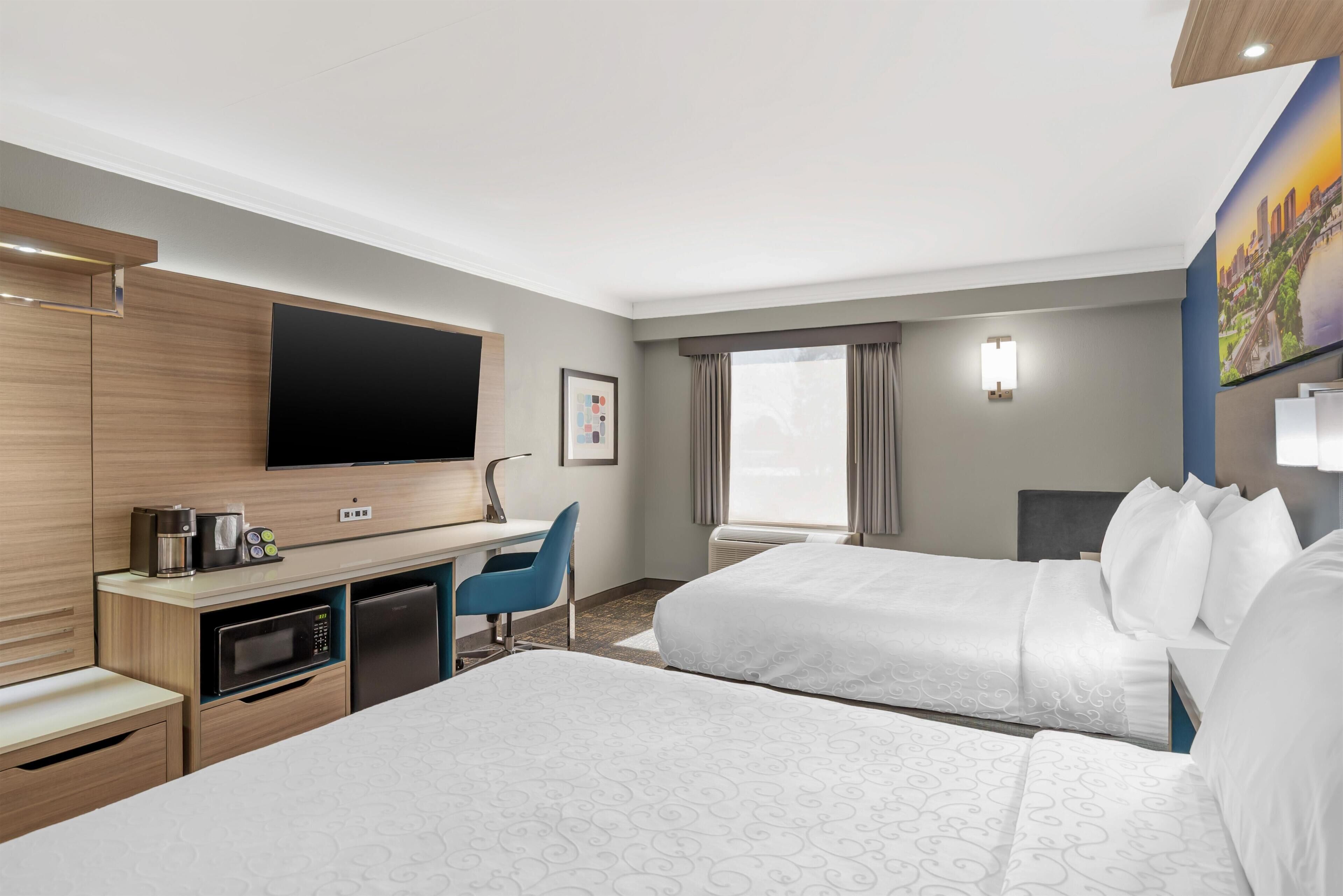 Habitación de hotel con televisión de pantalla plana, escritorio con cafetera, microondas, refrigerador, dos camas, una silla azul y un ventanal con cortinas.
