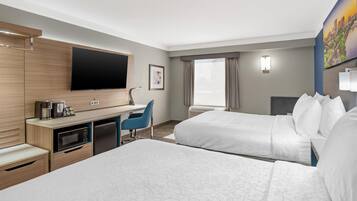 Ein Hotelzimmer mit Flachbildfernseher, Schreibtisch mit Kaffeemaschine, Mikrowelle, Kühlschrank, zwei Betten, blauem Sessel und großem Fenster mit Vorhängen.