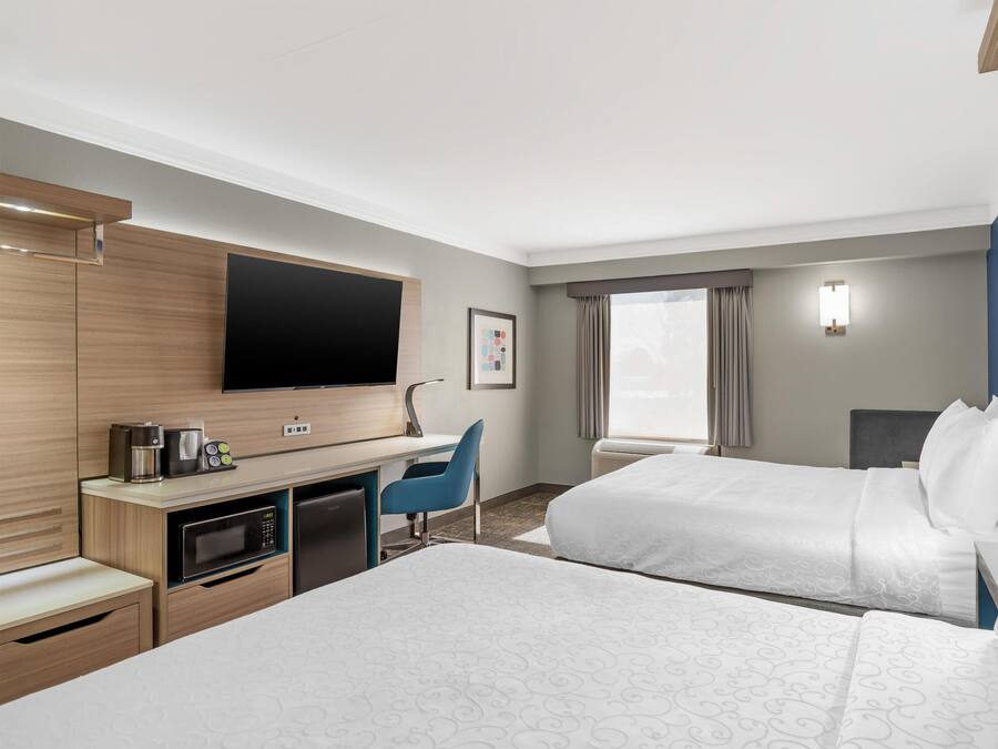 Ein Hotelzimmer mit Flachbildfernseher, Schreibtisch mit Kaffeemaschine, Mikrowelle, Kühlschrank, zwei Betten, blauem Sessel und großem Fenster mit Vorhängen.