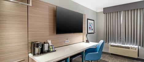 Une chambre d’hôtel moderne équipée d’un téléviseur à écran plat, d’une machine à café, d’un four à micro-ondes, d’un mini-réfrigérateur, d’une chaise bleue et d’une grande fenêtre avec des rideaux.