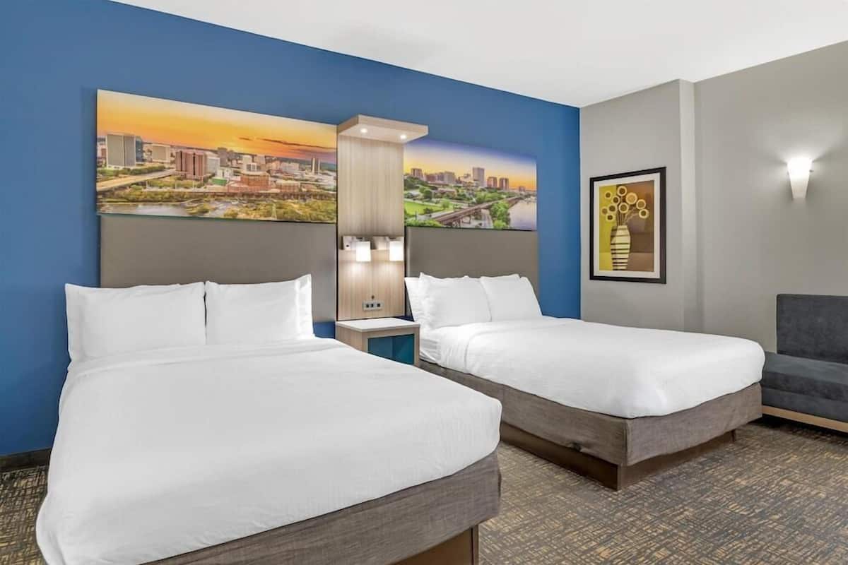 Habitación de hotel con dos camas, una mesita de noche, una lámpara y una pintura de un paisaje urbano en la pared.