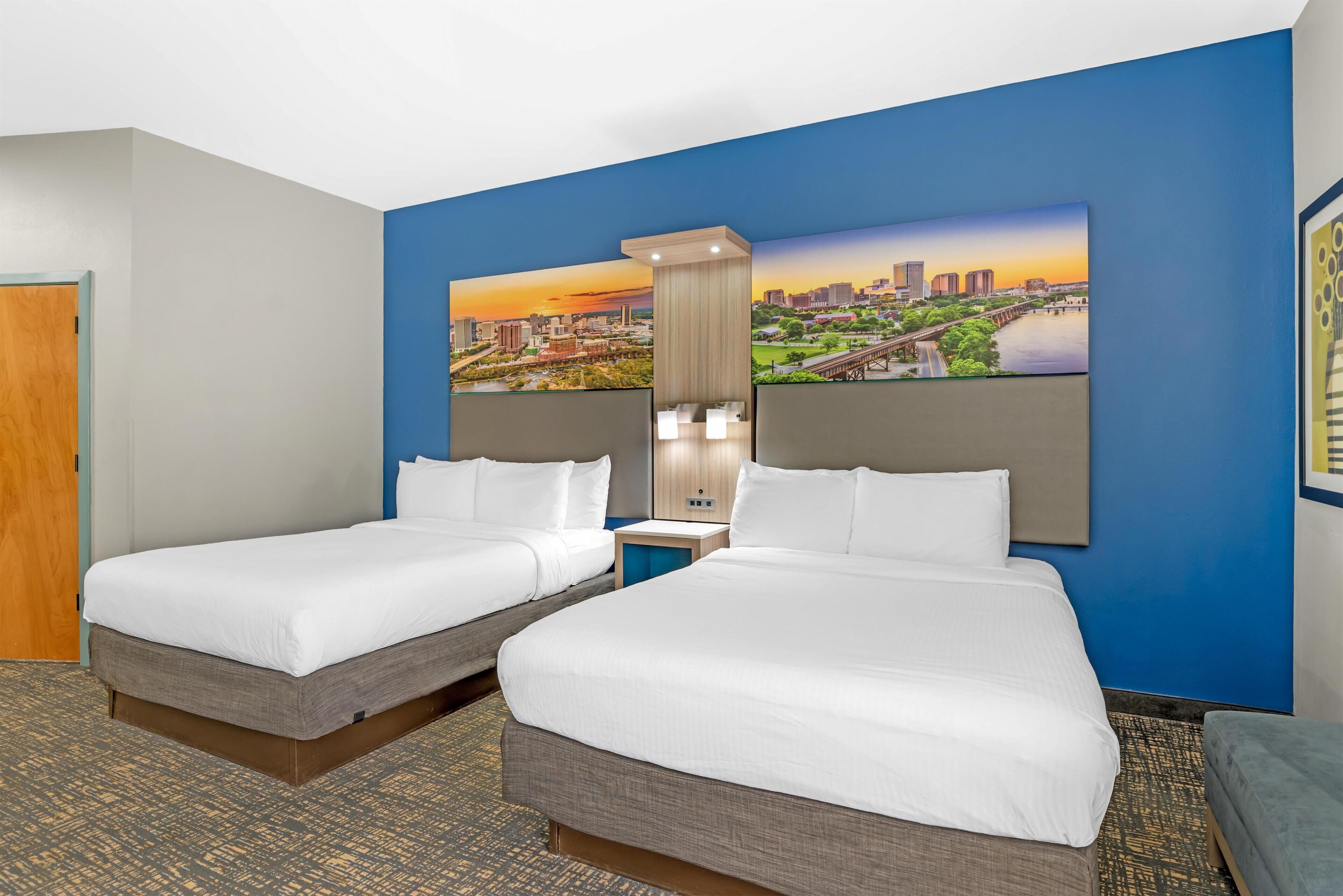 A hotel room with two beds, a cityscape painting on the wall, a nightstand, and a carpeted floor.