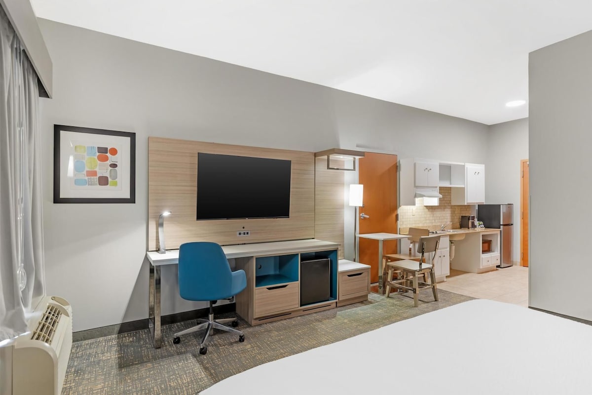 Une chambre d’hôtel équipée d’un bureau, d’une chaise, d’une télévision, d’une kitchenette et d’un tableau encadré au mur.