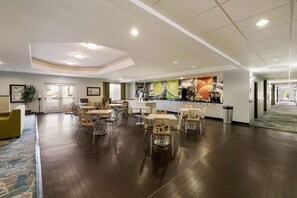 A spacious hotel lobby with multiple dining tables and chairs, a reception desk, and a large mural on the wall.