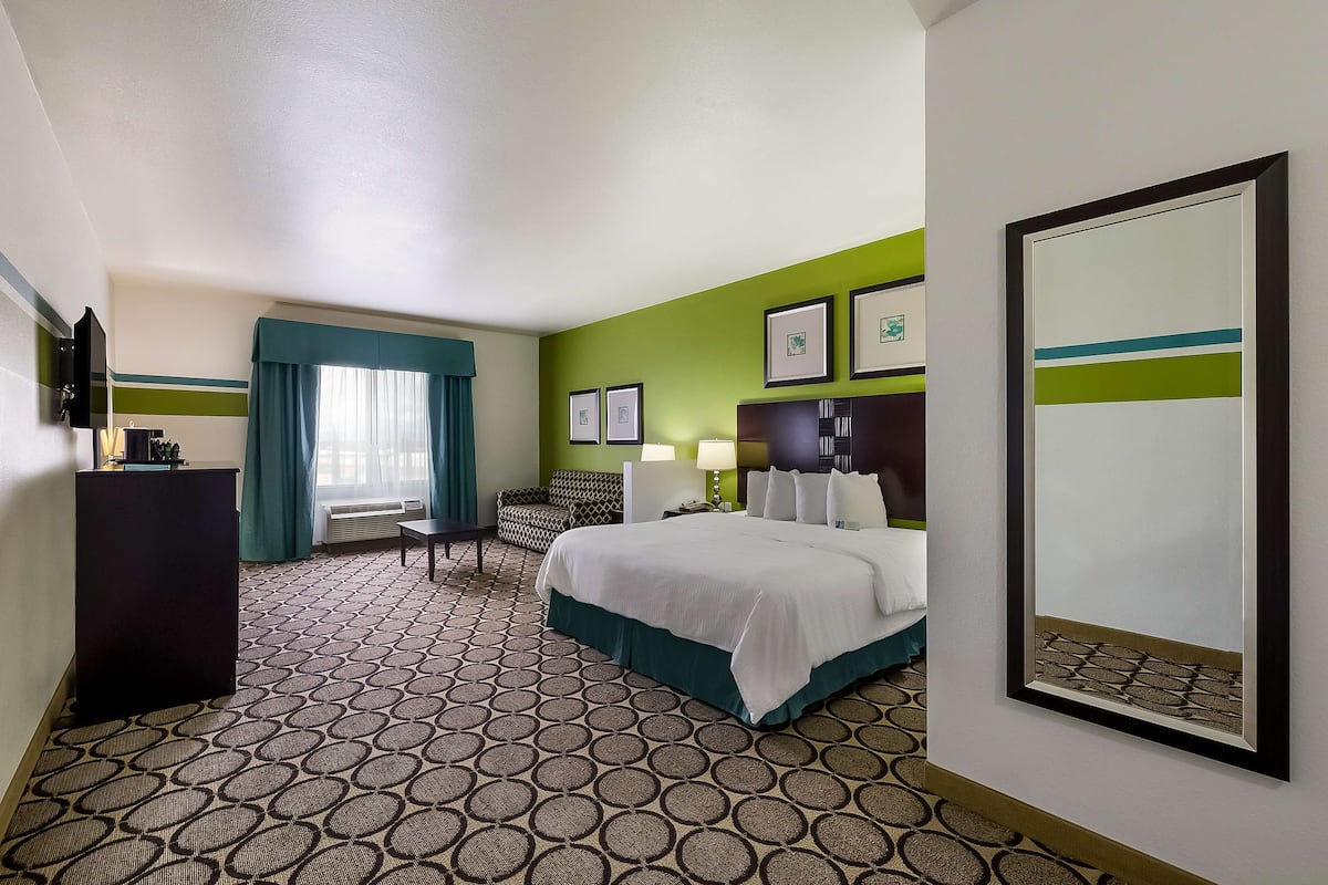 A hotel room with a bed, a desk, a chair, a window with curtains, a wall-mounted TV, and framed pictures.