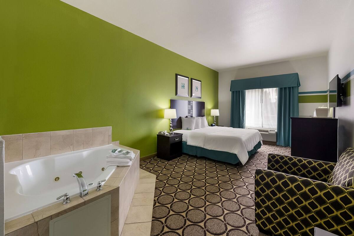 A hotel room with a large bathtub, a bed with a blue headboard, a desk, and a patterned carpet.