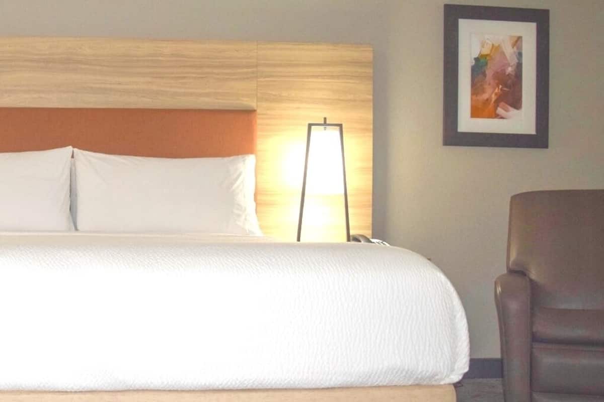 A hotel room with a large bed, a headboard, a standing lamp, a framed picture on the wall, and a brown leather chair.