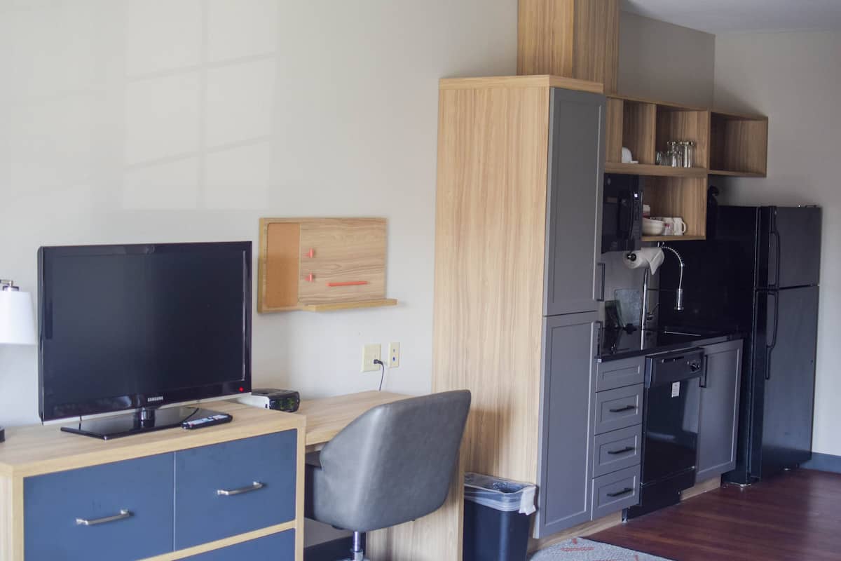A compact hotel room with a TV, wooden desk, chair, and kitchenette.
