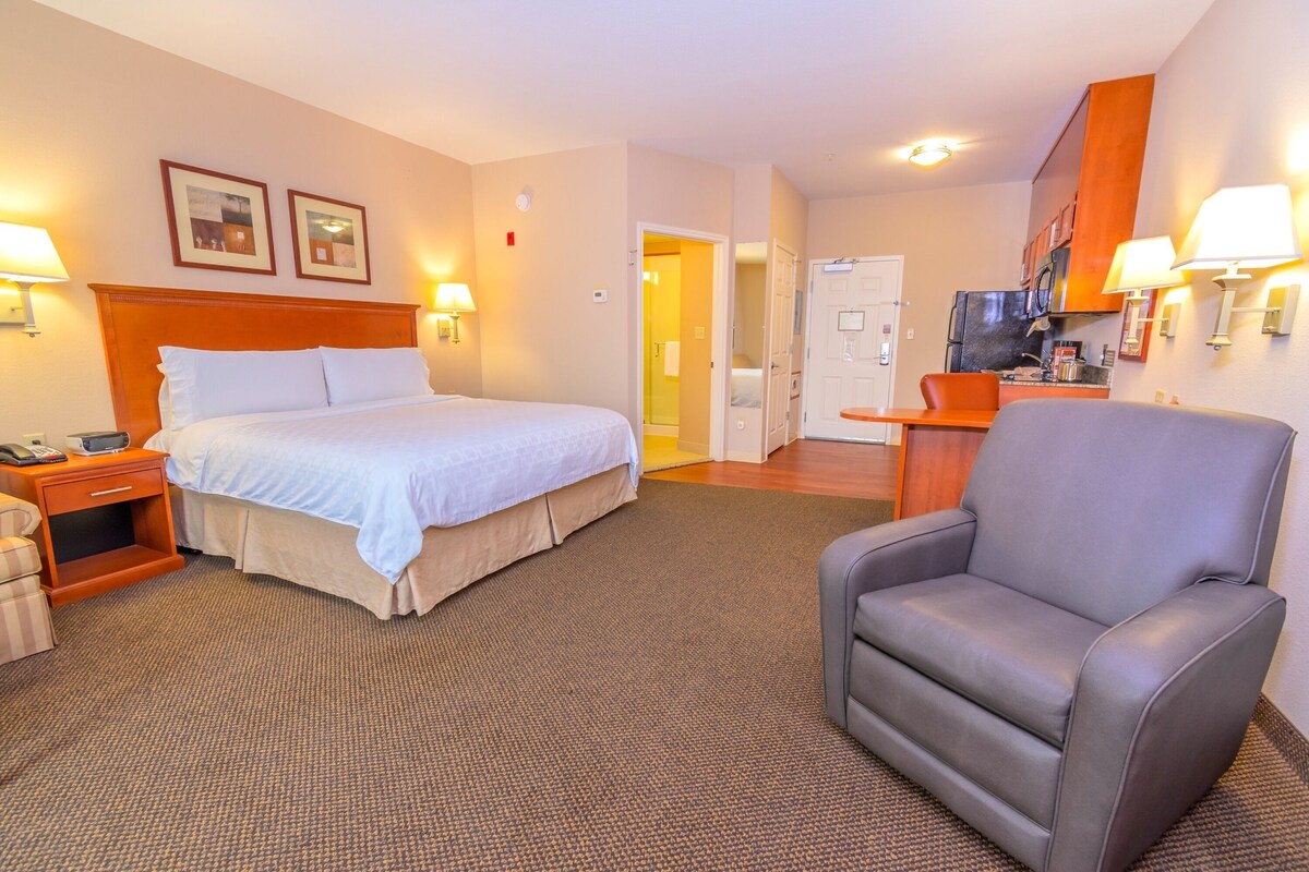 A hotel room with a bed, a chair, a nightstand, a desk, and a kitchenette.
