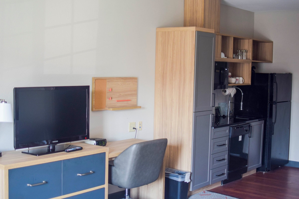 A compact hotel room with a TV, wooden desk, chair, and kitchenette.