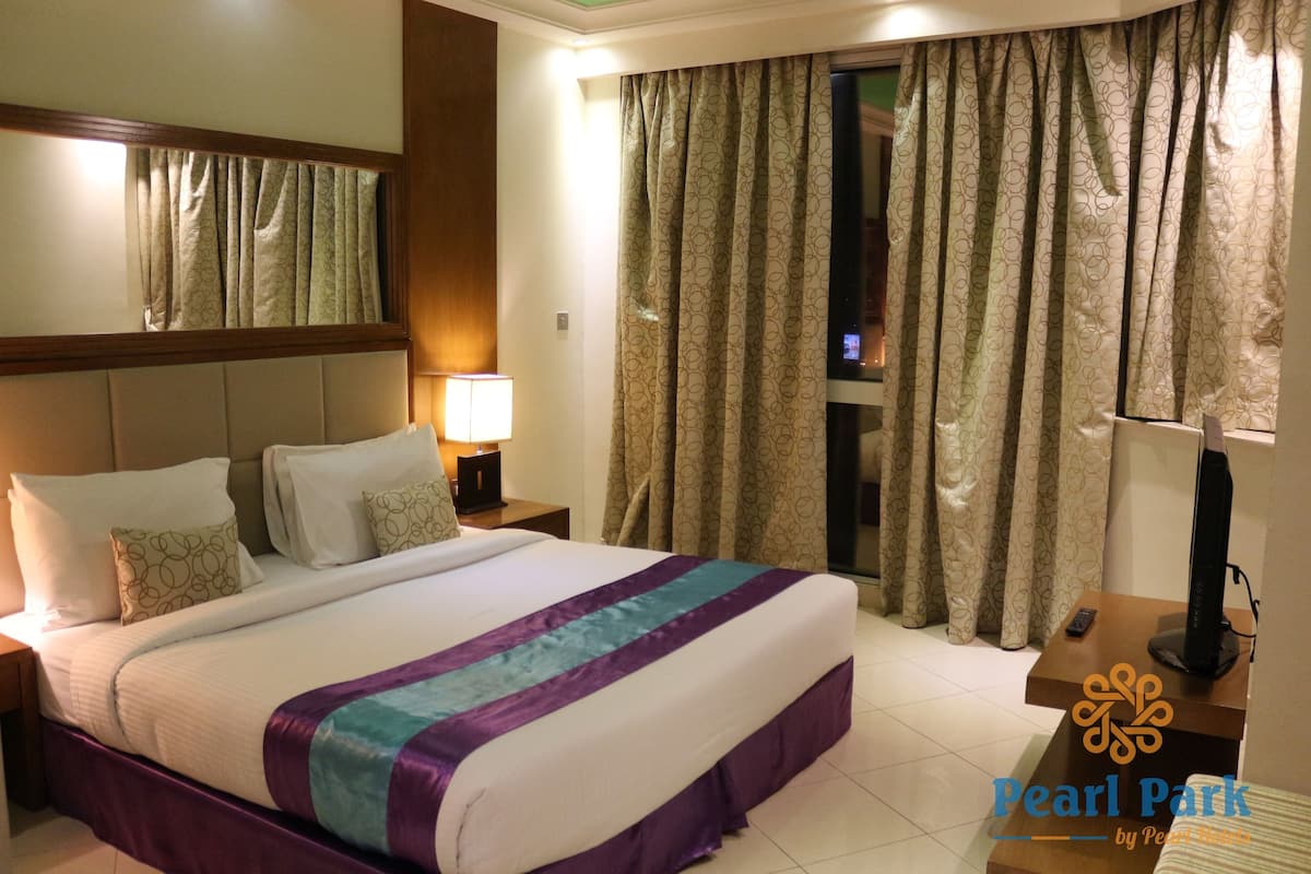 Una habitación de hotel con una cama grande, una mesita de noche con lámpara, un televisor sobre un mueble bajo y cortinas en la ventana.
