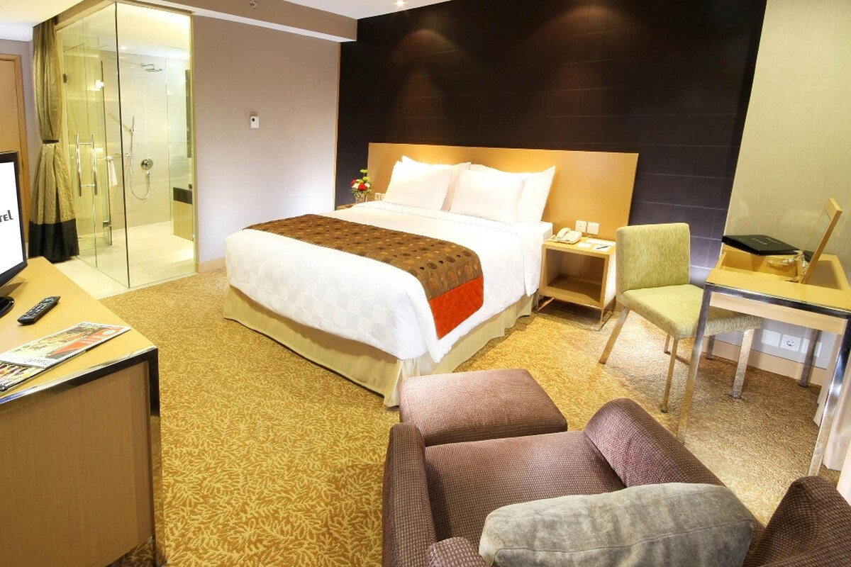 A hotel room with a large bed, a desk with a computer, a chair, and a glass-enclosed shower area.