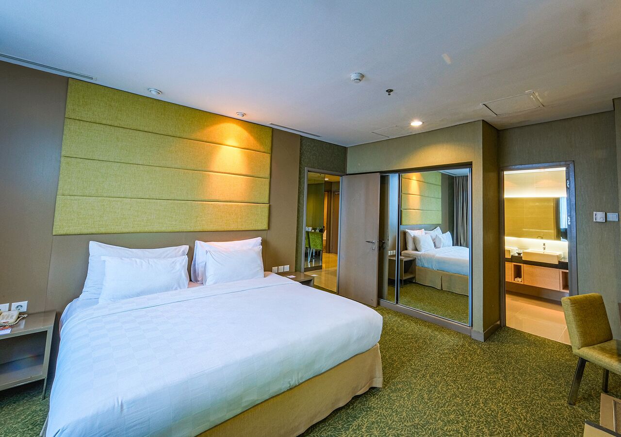 Habitación de hotel con una cama grande, una mesita de noche, una silla, un espejo y un baño visible a través de una puerta abierta.