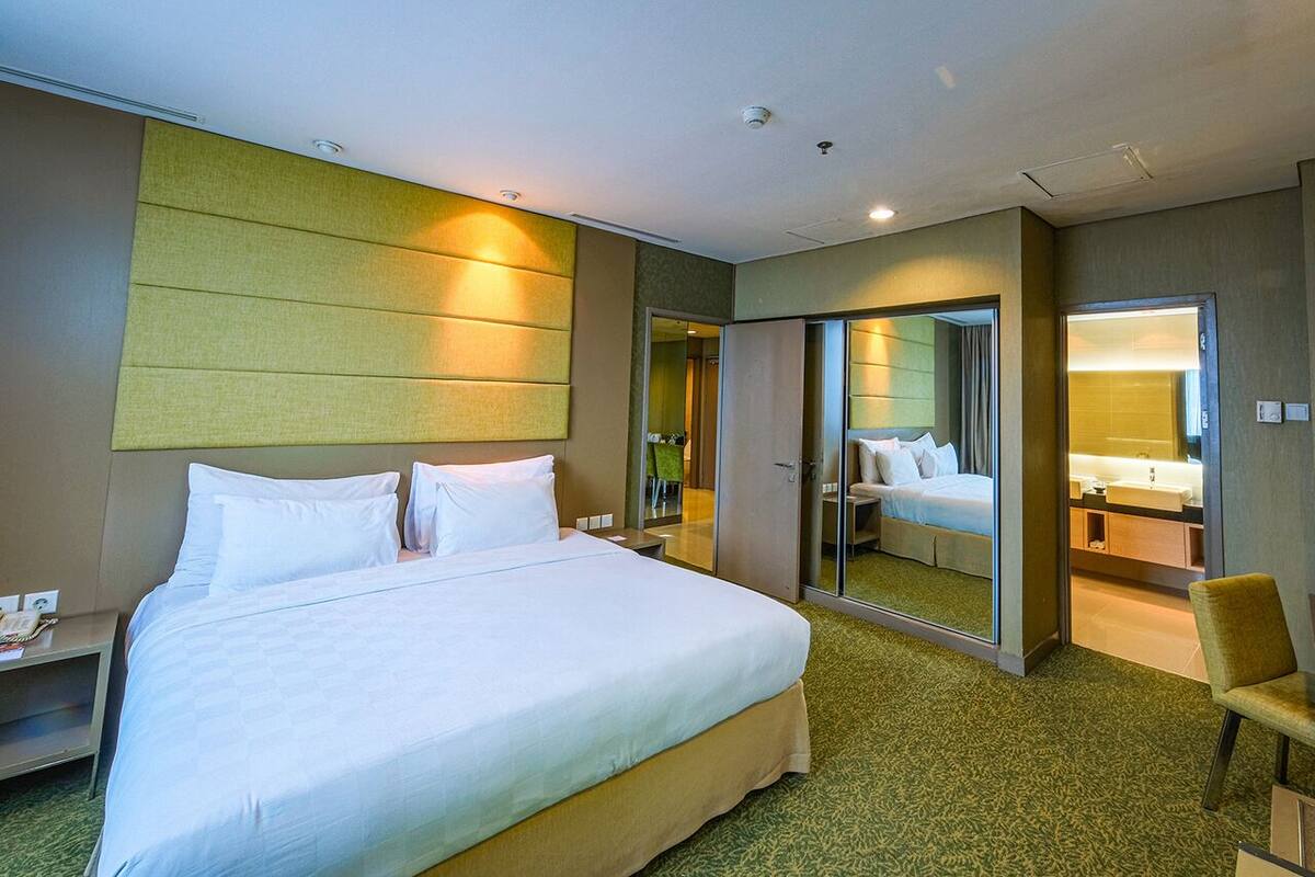 Ein Hotelzimmer mit einem großen Bett, einem Nachttisch, einem Sessel, einem Spiegel und einem durch eine offene Tür einsehbaren Badezimmer.