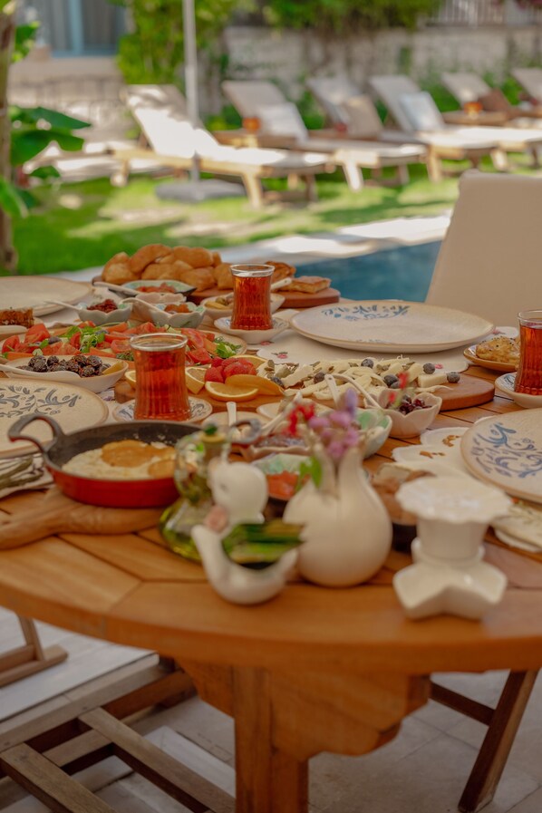 A round wooden table with a variety of dishes and drinks, surrounded by lounge chairs and a swimming pool in the background. - Villa Fora Hotel (Cesme)