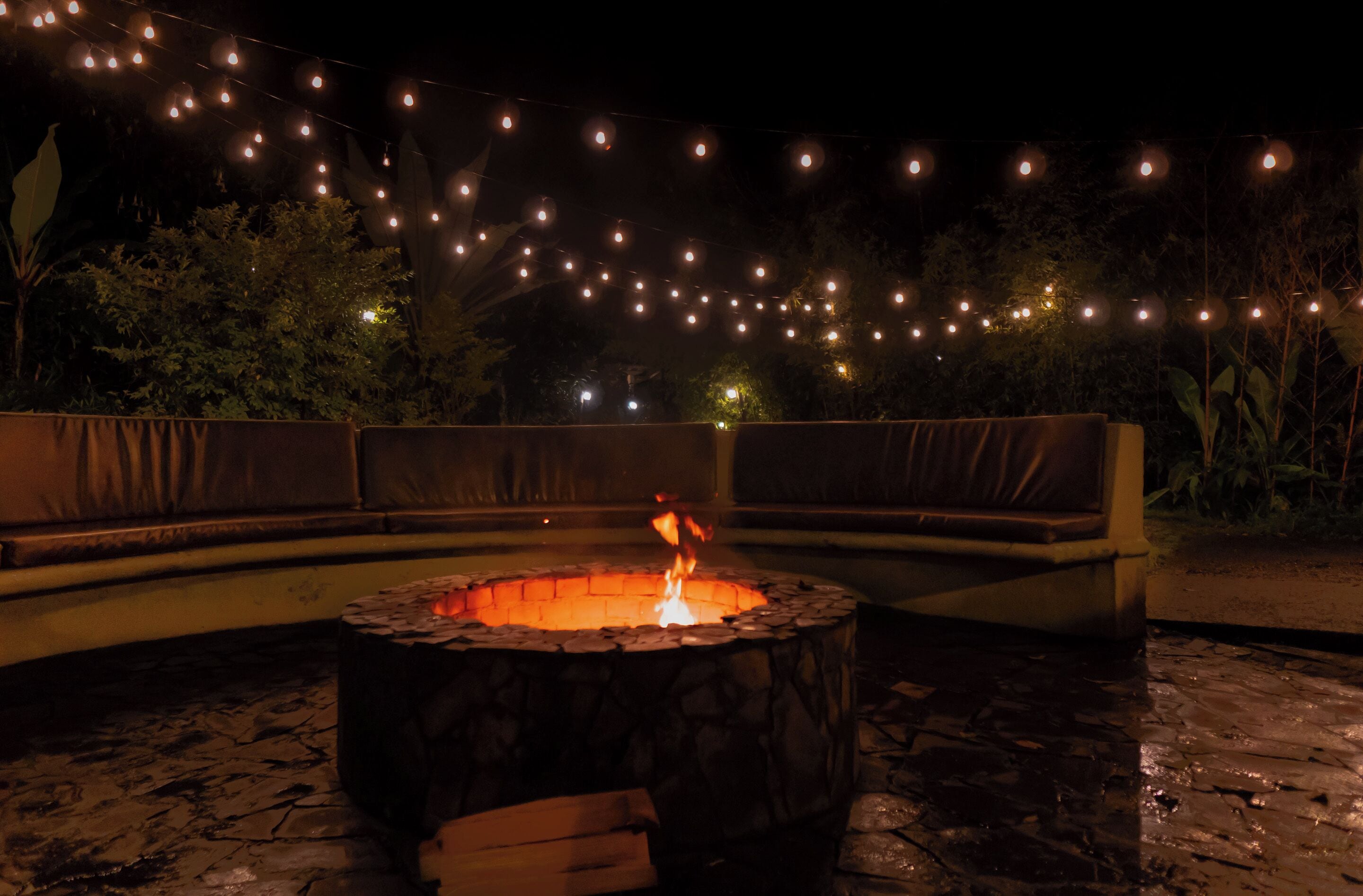 Un espacio al aire libre con una chimenea circular, rodeada de suelo de piedra y luces colgantes de fondo.