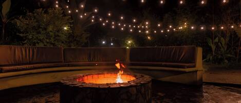 An outdoor seating area with a circular fire pit, surrounded by stone flooring, and string lights in the background.