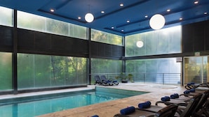A modern indoor swimming pool with clear glass walls, lounge chairs, and a ceiling with recessed lighting.
