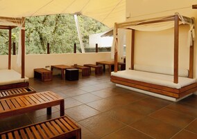 Un espacio exterior techado con mobiliario de madera, incluyendo bancos y una cama para el día, bajo un dosel.