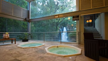 Relax in a serene indoor space with two hot tubs, lush greenery, and a soothing waterfall view.