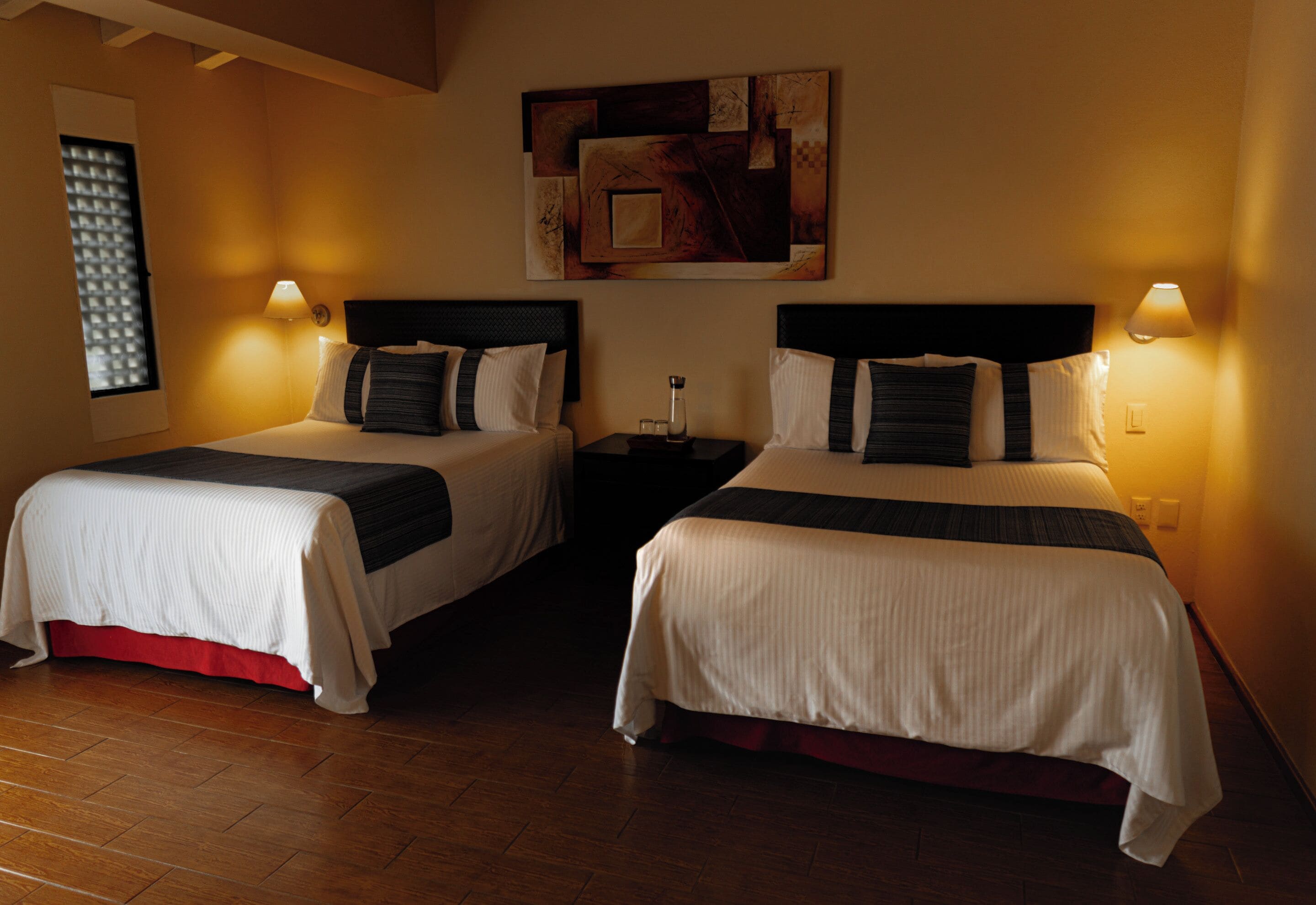A hotel room with two beds, each with a headboard, bedside lamps, and a small table between them.