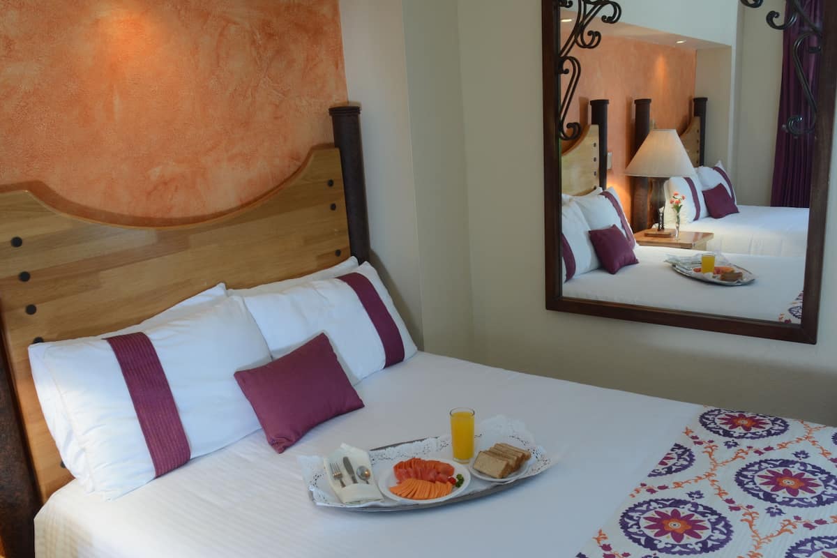 A hotel room with a wooden headboard, a bed with white and purple bedding, a tray with breakfast including toast, fruit, and juice, and a mirror reflecting the room.