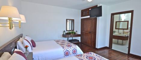 Habitación de hotel con dos camas, ventilador de techo, televisión, armario de madera, espejo y un pequeño escritorio con una planta.