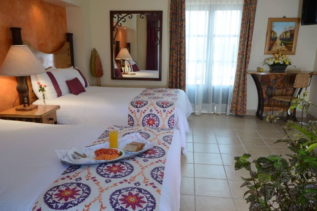 A hotel room with two beds, a dining table with breakfast, a mirror, a desk, and a plant.