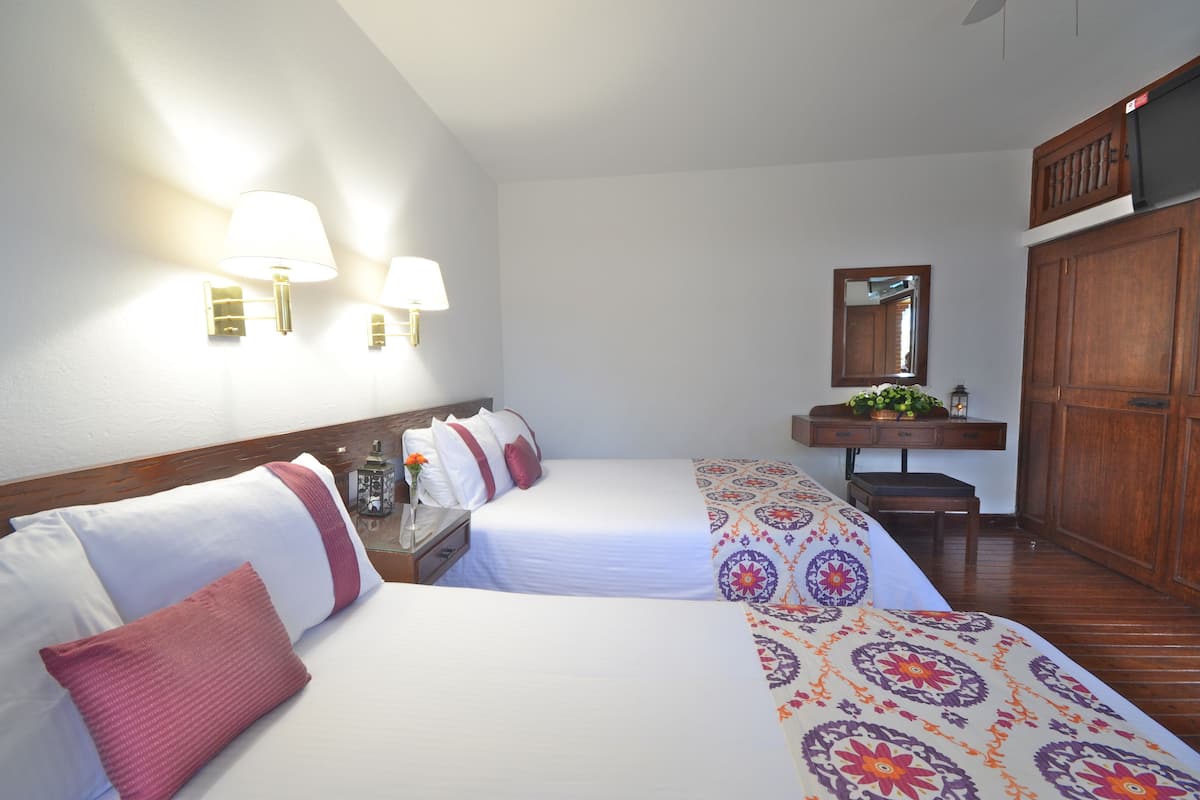 A hotel room with two beds, a wooden wardrobe, a bedside table with a lamp, and a mirror on the wall.