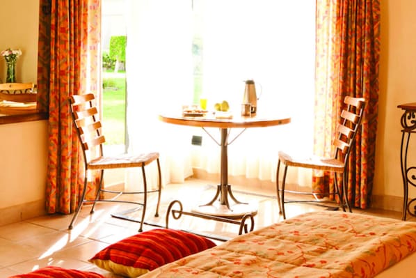 A room with a bed, a round table with chairs, and a view of a garden through a window.