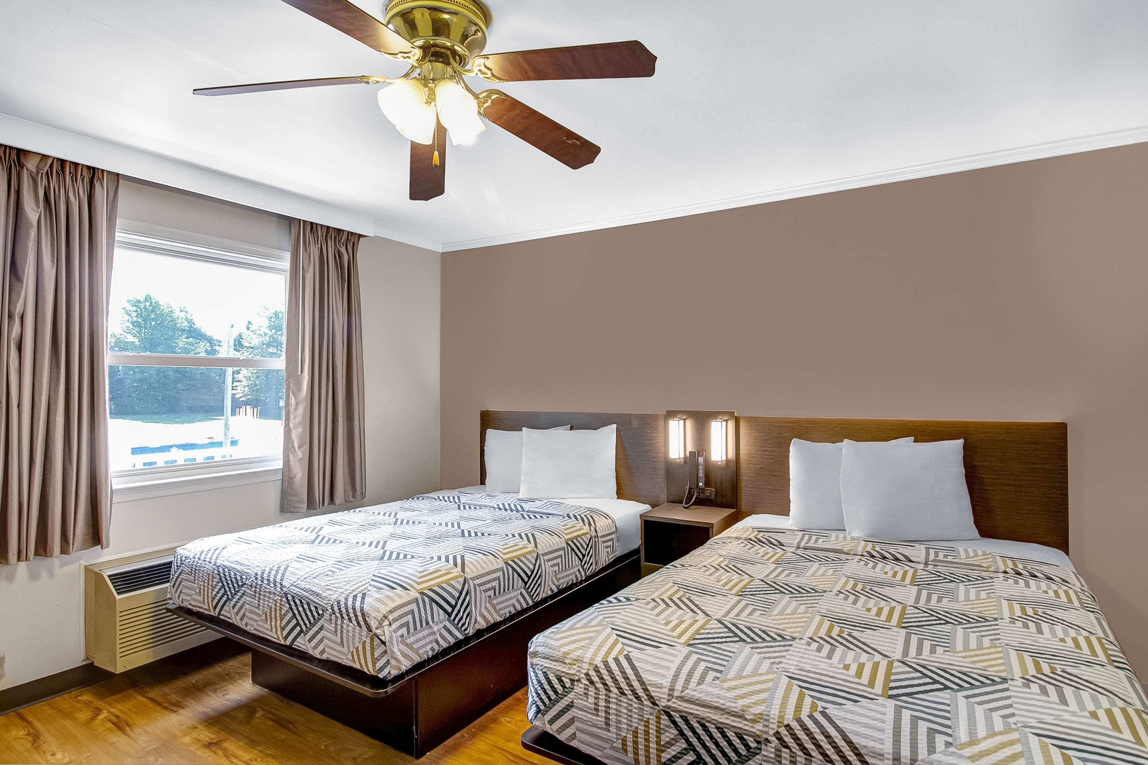 A hotel room with two beds, a ceiling fan, a window with curtains, and a view of the outside.