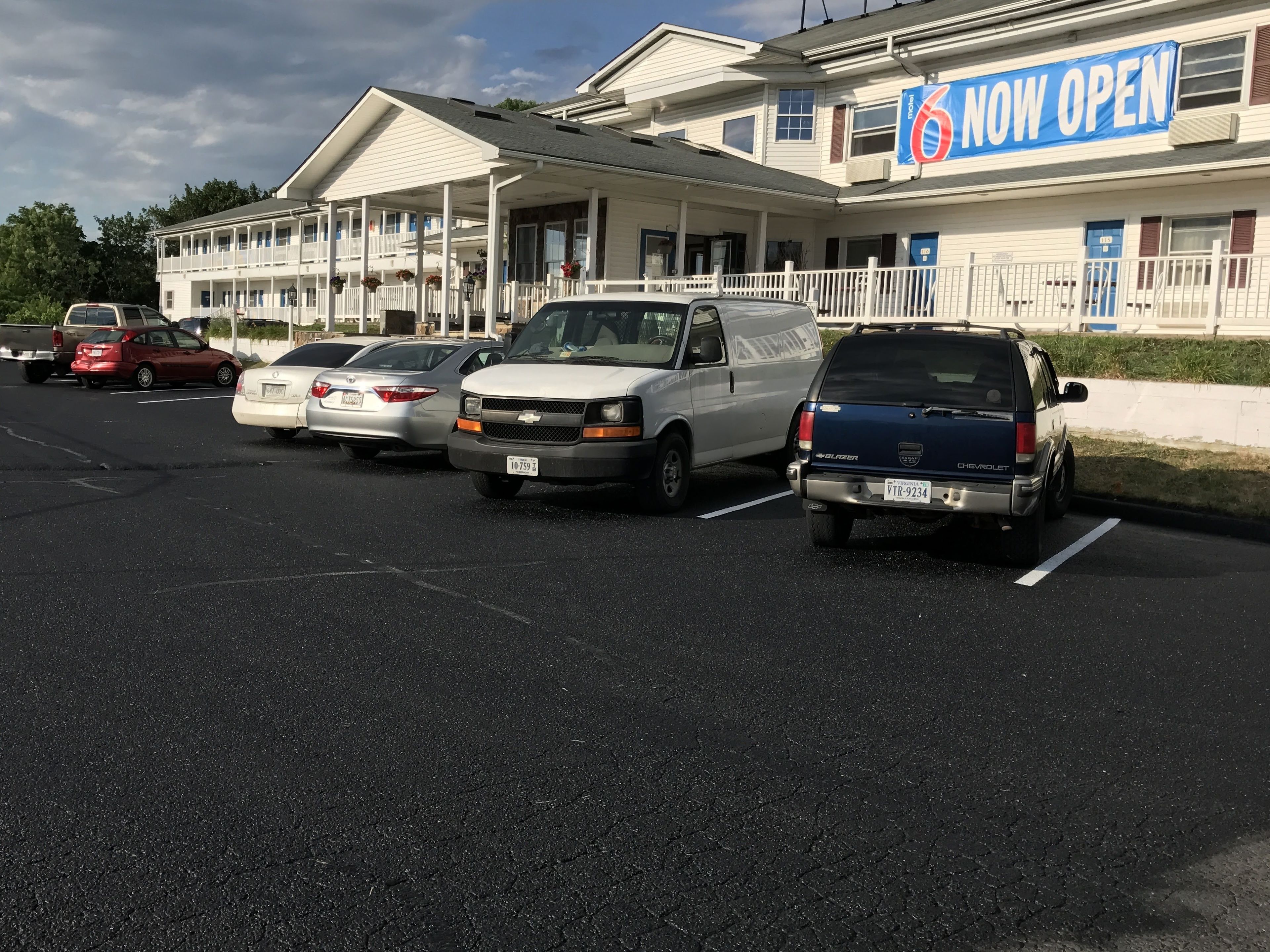 Un motel con un cartel de 'Abierto ya', que muestra un estacionamiento con varios vehículos, incluyendo una camioneta blanca y un SUV azul.
