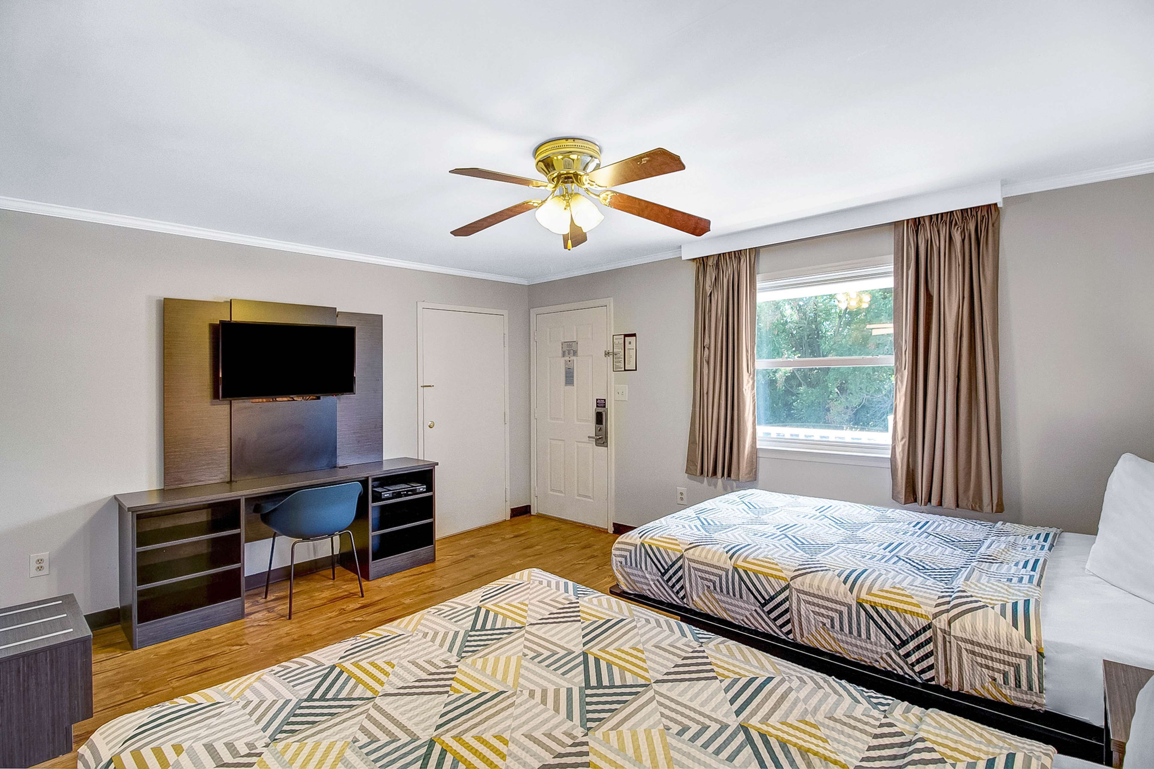 Ein Hotelzimmer mit zwei Betten, einem Schreibtisch, einem Fernseher, einem Deckenventilator und einem Fenster mit Vorhängen.