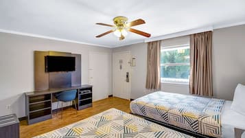 Ein Hotelzimmer mit zwei Betten, einem Schreibtisch, einem Fernseher, einem Deckenventilator und einem Fenster mit Vorhängen.