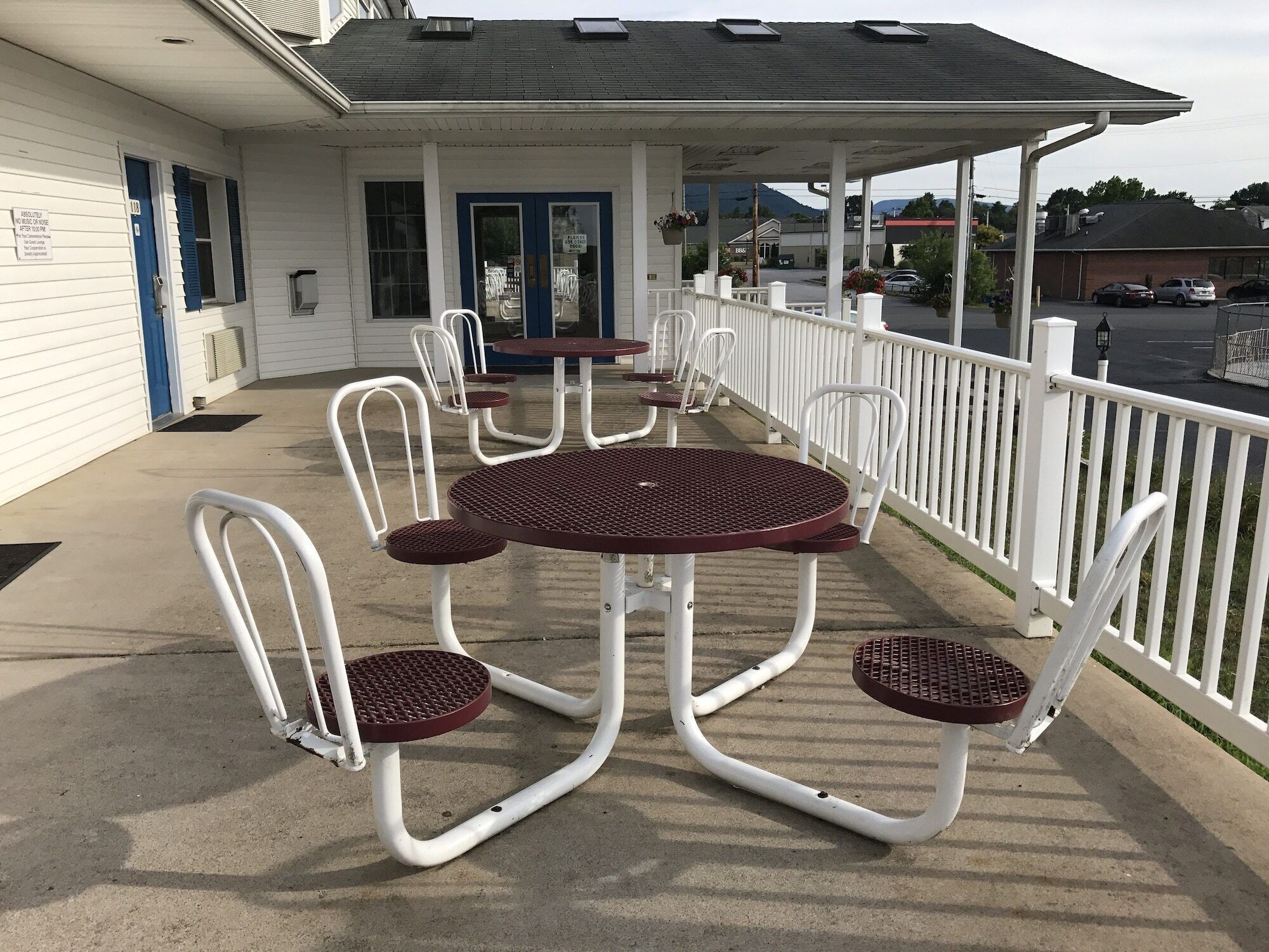A covered outdoor seating area with white metal chairs and tables, a blue door, and a white building with a dark roof.