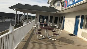 A covered outdoor seating area with tables and chairs, a white fence, and a building with a 'NOW OPEN' sign.
