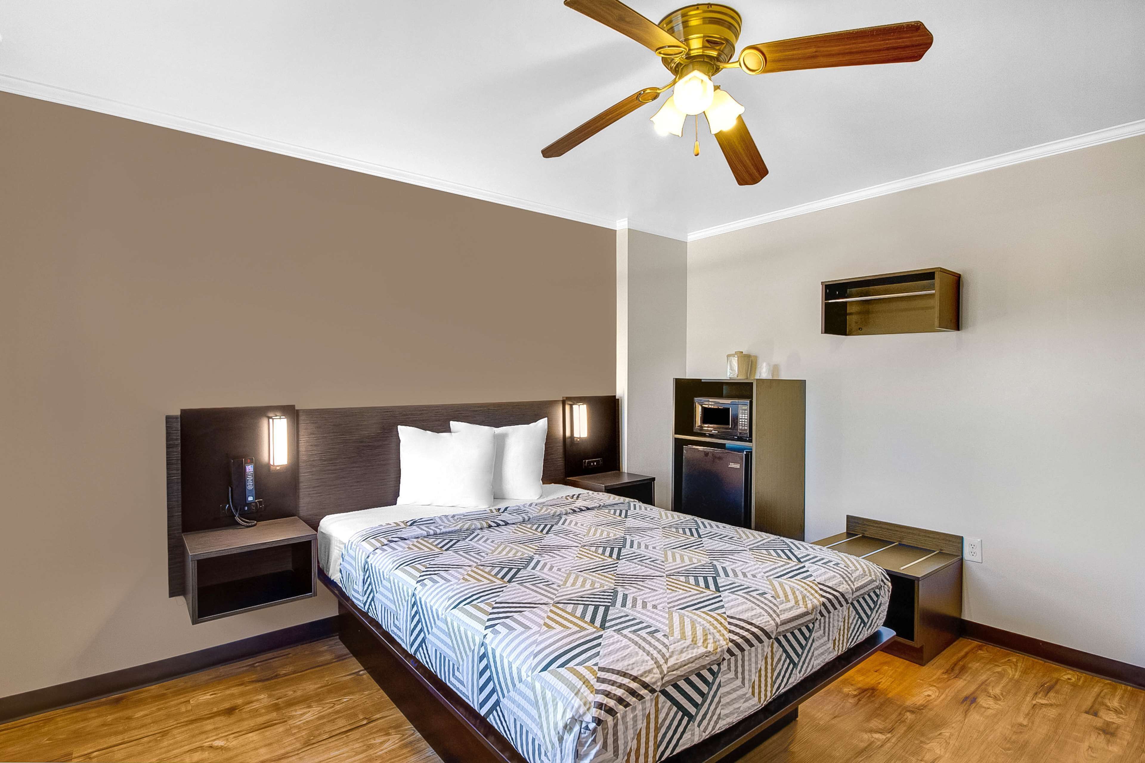 Una habitación de hotel con cama, mesita de noche, escritorio, ventilador de techo y televisor montado en la pared.