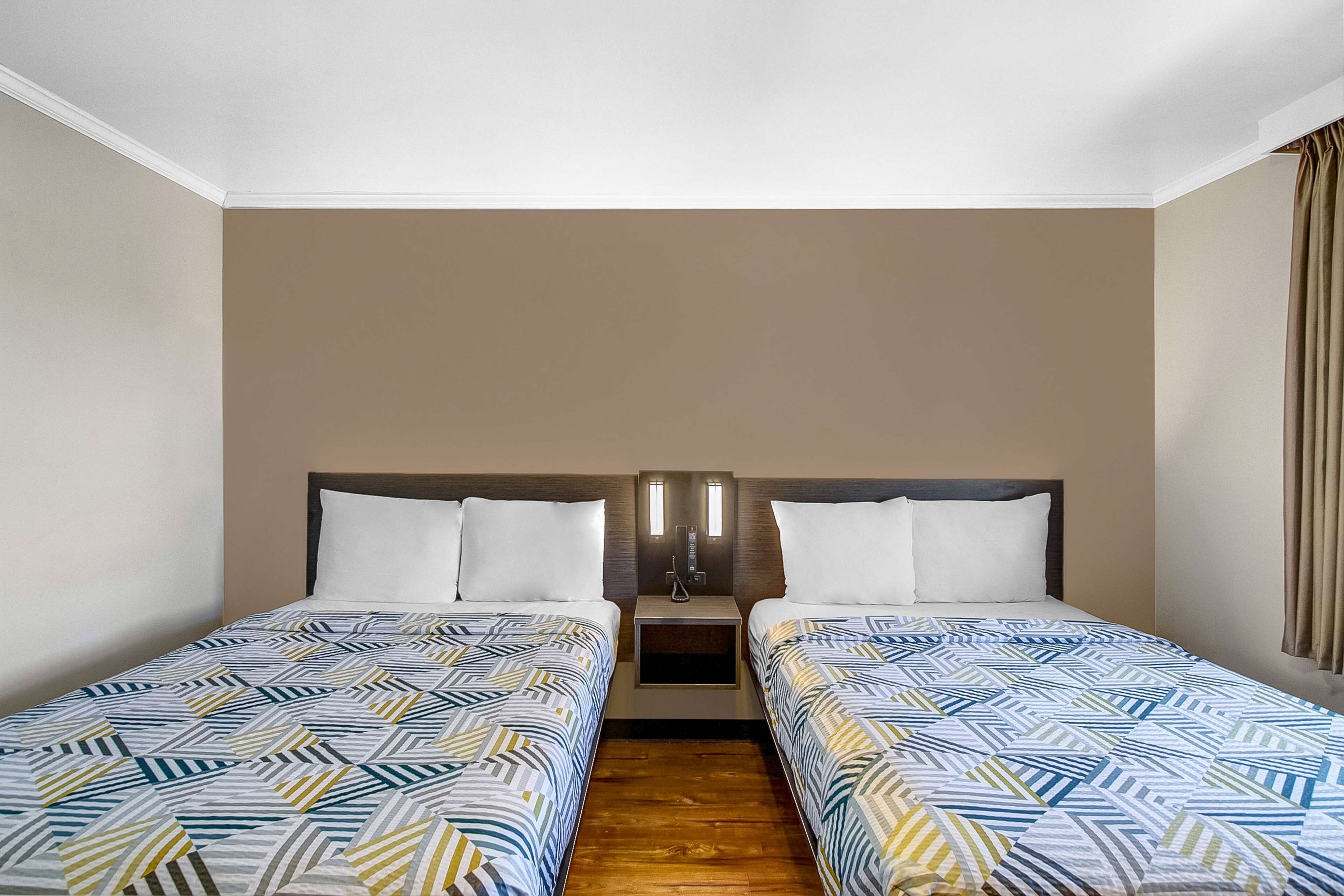 Habitación de hotel con dos camas individuales, piso de madera, mesita de noche con lámpara y pared lisa.