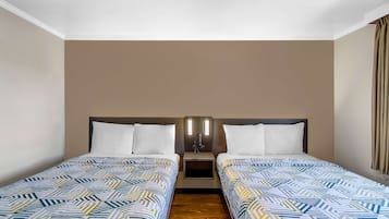 Habitación de hotel con dos camas individuales, piso de madera, mesita de noche con lámpara y pared lisa.