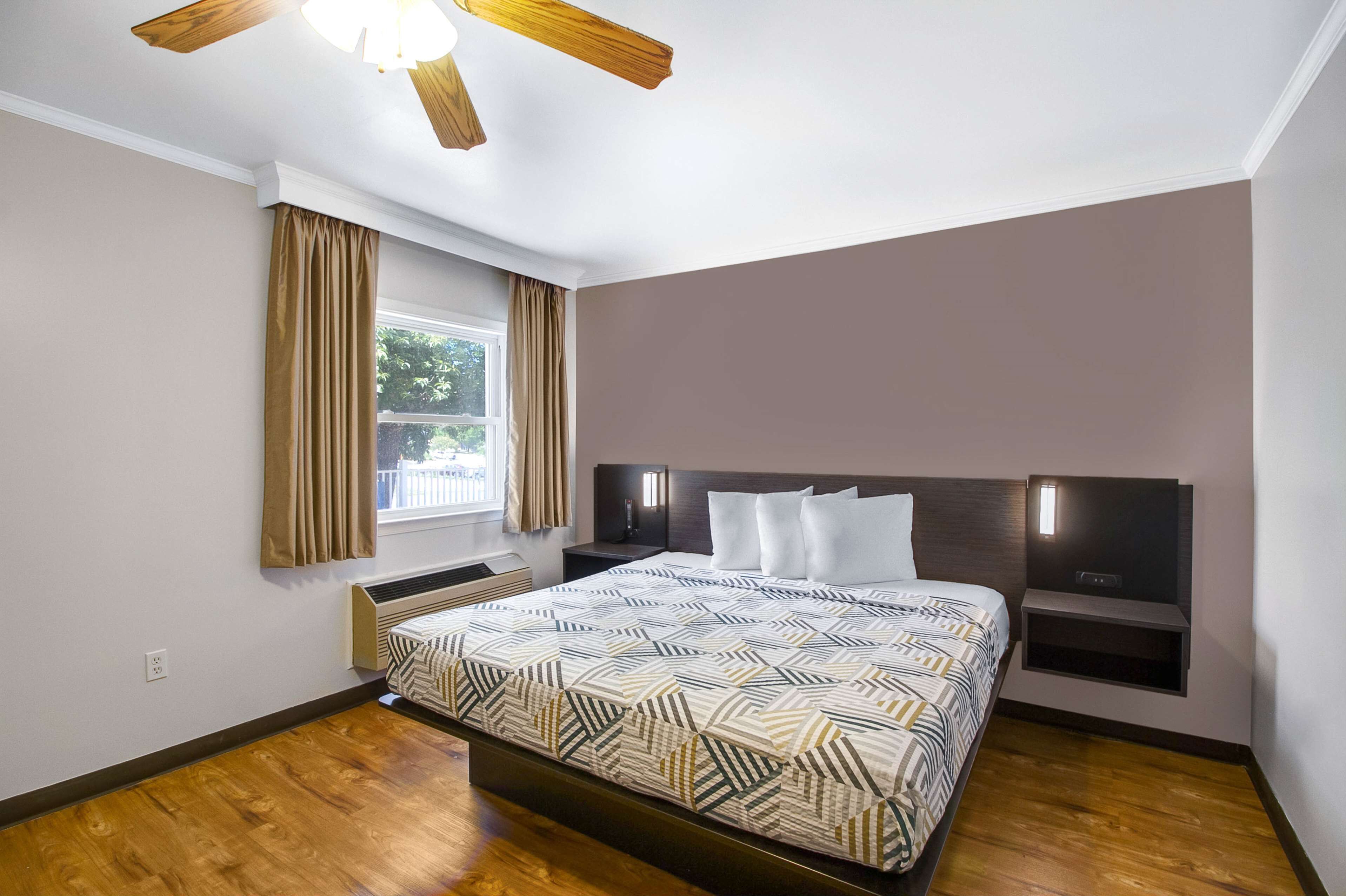 Un dormitorio con cama, ventilador de techo, ventana con cortinas, mesita de noche y televisor montado en la pared.