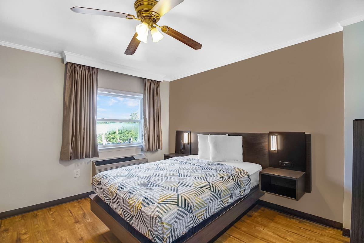 A hotel room with a bed, a ceiling fan, a window with curtains, a nightstand, and a wall-mounted shelf.
