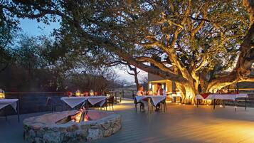 Un espace de restauration en plein air avec des tables disposées sous un grand arbre, entouré de murs en pierre et agrémenté d’un foyer.