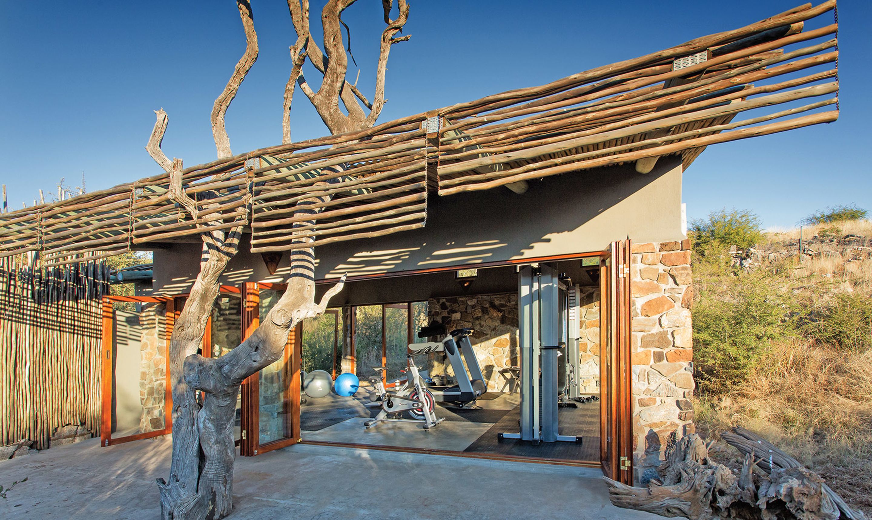 Un bâtiment moderne avec une façade en pierre, une pergola en bois et des équipements de musculation à l’intérieur.