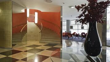 Un vestíbulo con piso a cuadros, una escalera con barandilla curva y un jarrón grande con flores rojas.