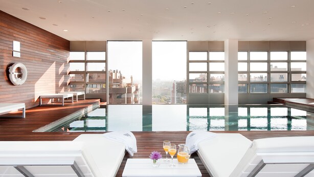 Un área de piscina interior moderna con una terraza de madera, sillones de descanso y una mesa con bebidas y una flor.
