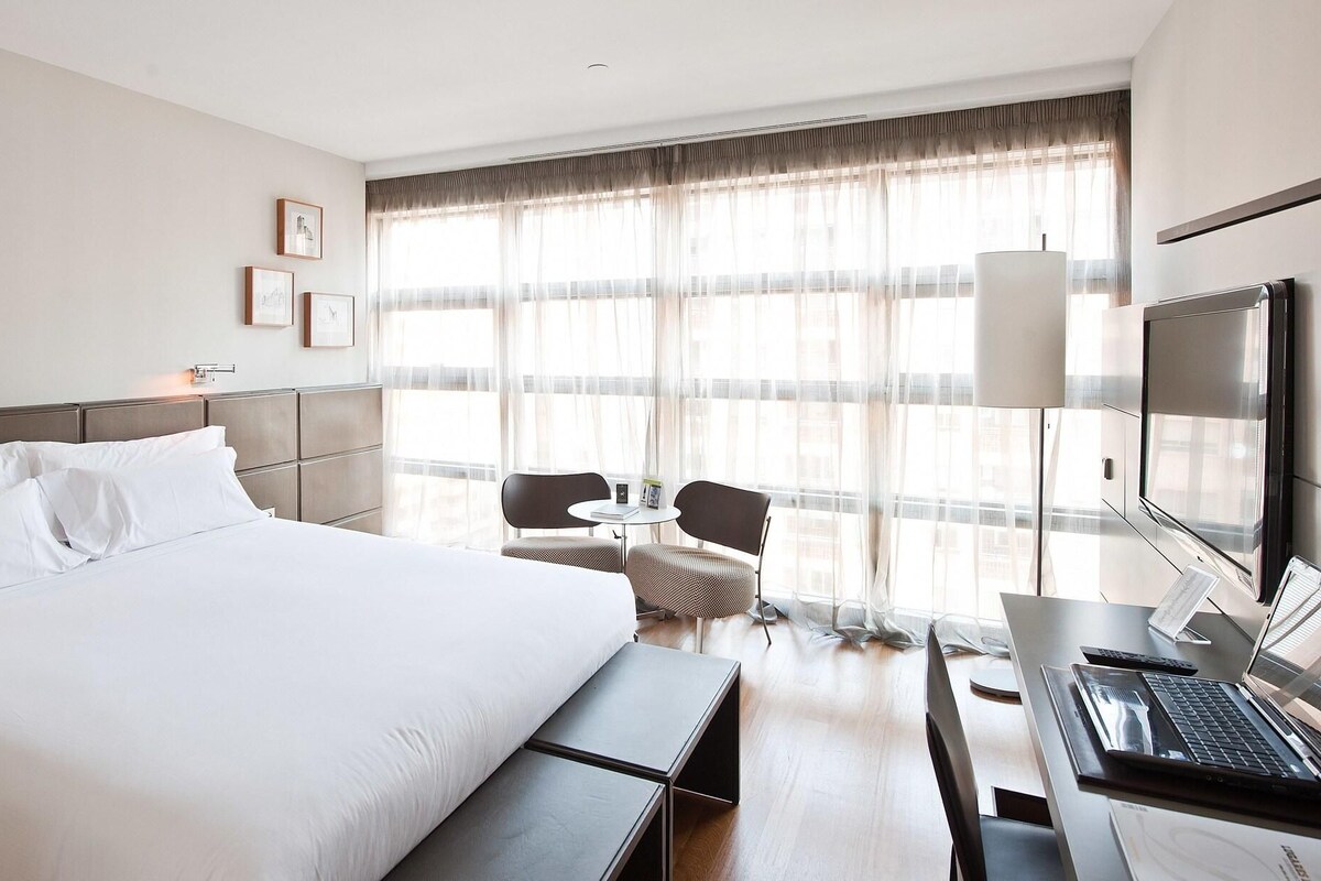 Habitación de hotel con una cama grande, un escritorio con una computadora portátil, un televisor y una ventana con cortinas translúcidas.