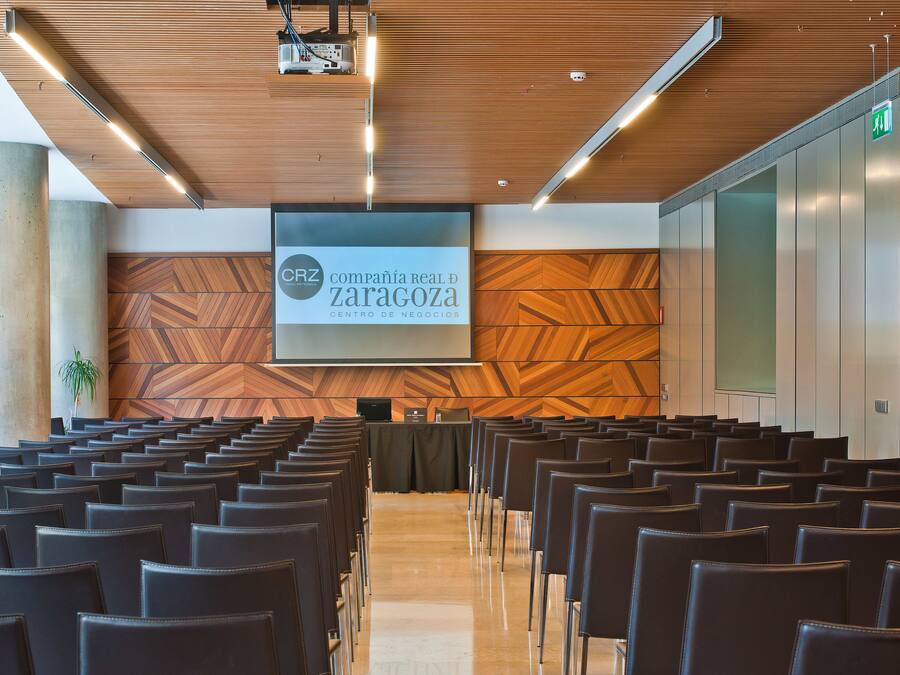A conference room with rows of chairs, a projection screen, and a wooden wall with a geometric pattern.