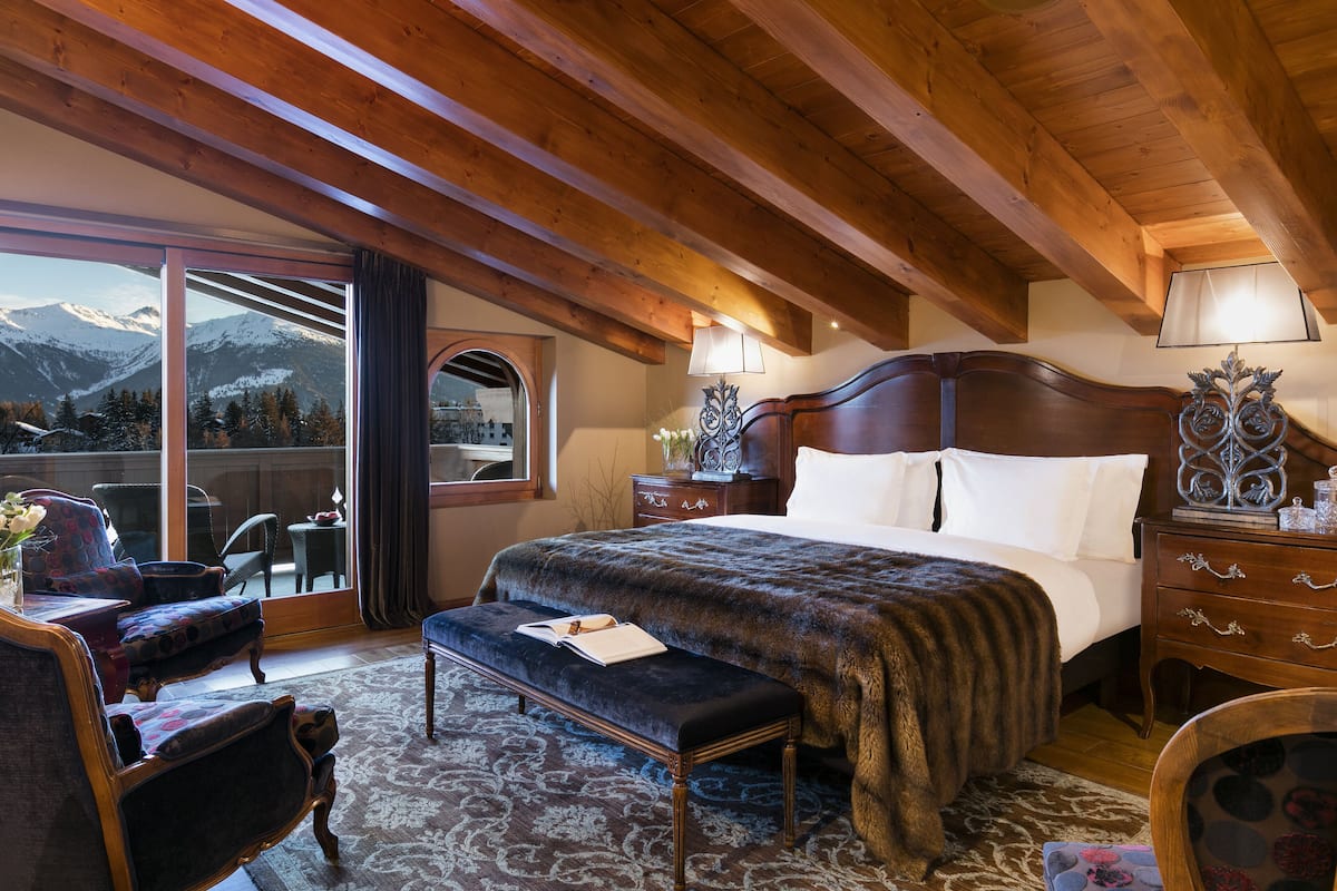 Ein Schlafzimmer mit einem großen Bett, Holzbalken an der Decke, einer Bank, einem Sessel und Bergblick durch ein großes Fenster.