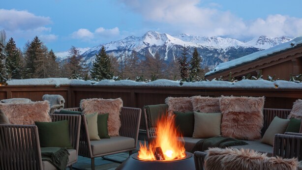 A cozy outdoor seating area with a fire feature, surrounded by snow-covered mountains and trees.