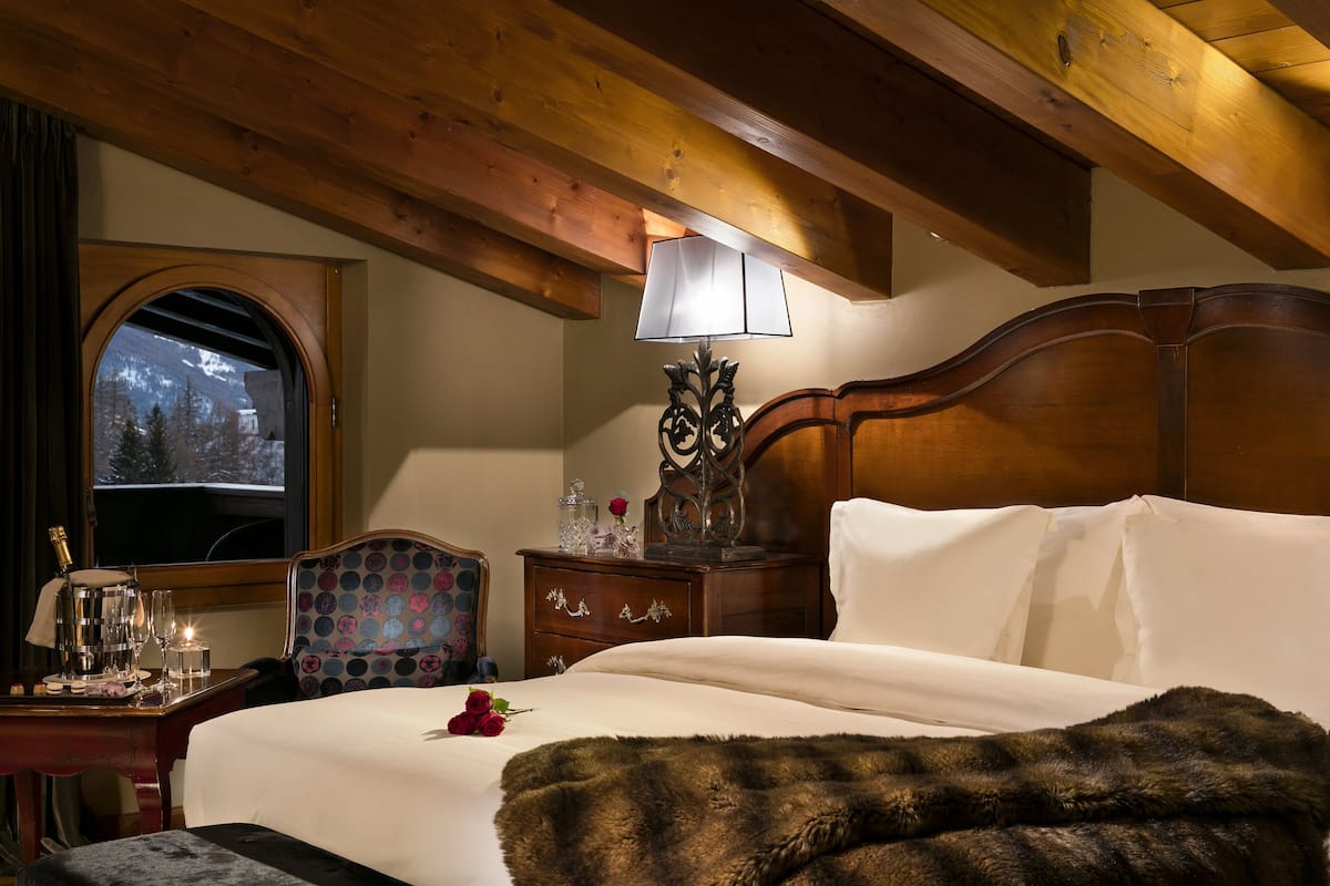 Ein Hotelzimmer mit Holzbalkendecke, einem großen Bett mit weißer Bettwäsche, einem Nachttisch aus Holz mit Lampe, einem Sessel und Blick auf schneebedeckte Berge durch ein Fenster.
